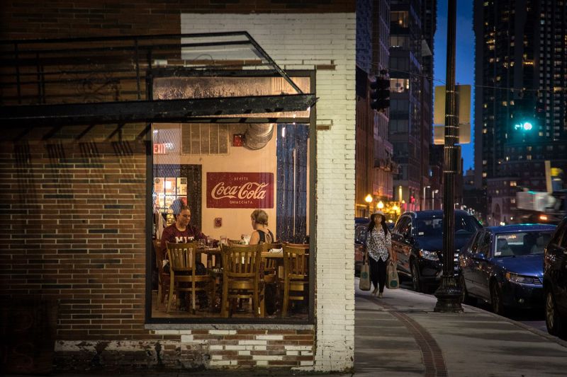 A nighttime street view of a corner restaurant with a large window showing two women dining, while a pedestrian carrying shopping bags walks down the adjacent city sidewalk.