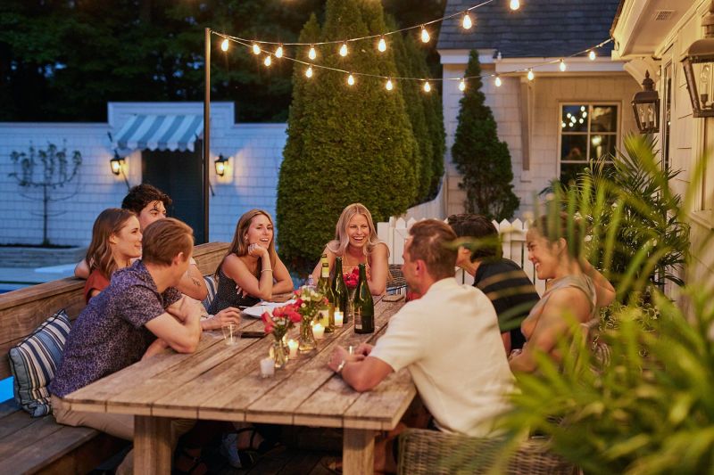 A group of smiling adults sitting around a rustic wooden table, talking and laughing during a backyard dinner party at twilight. The patio is illuminated by warm overhead string lights and small candles, with wine bottles and floral centerpieces on the ta